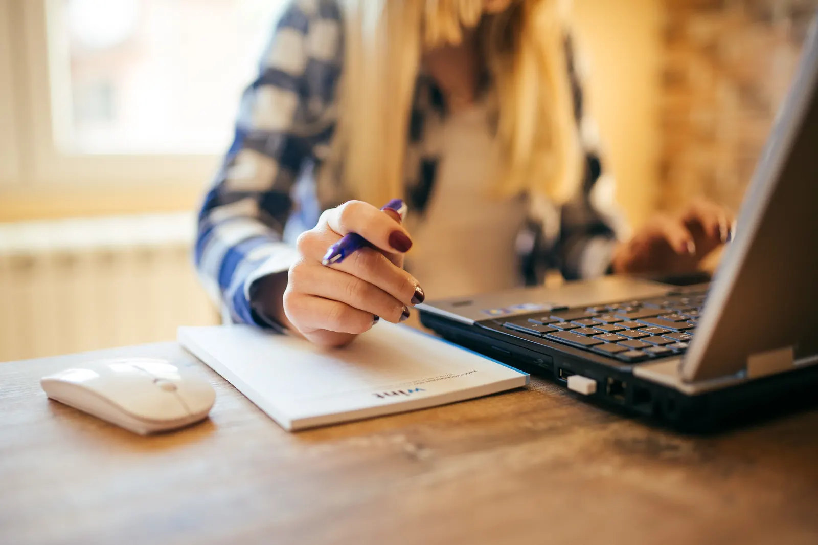 A freelancer making notes on a notepad next to a laptop at a wooden desk
